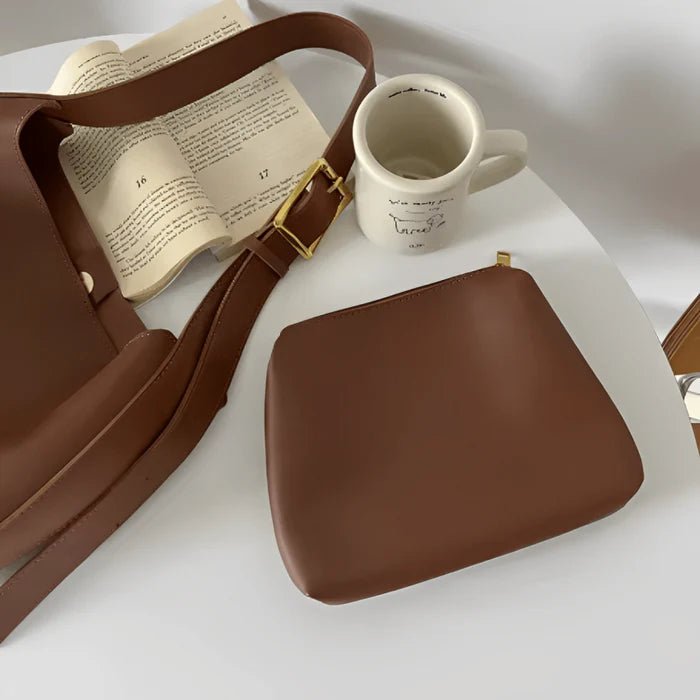 Bolso femenino Mavy de cuero sintético color marrón con asa ancha ajustable y hebilla dorada, acompañado de un bolsillo interior removible del mismo tono. El bolso está apoyado sobre una mesa blanca, al lado de un libro abierto y una taza blanca con ilustraciones y escritura en negro.